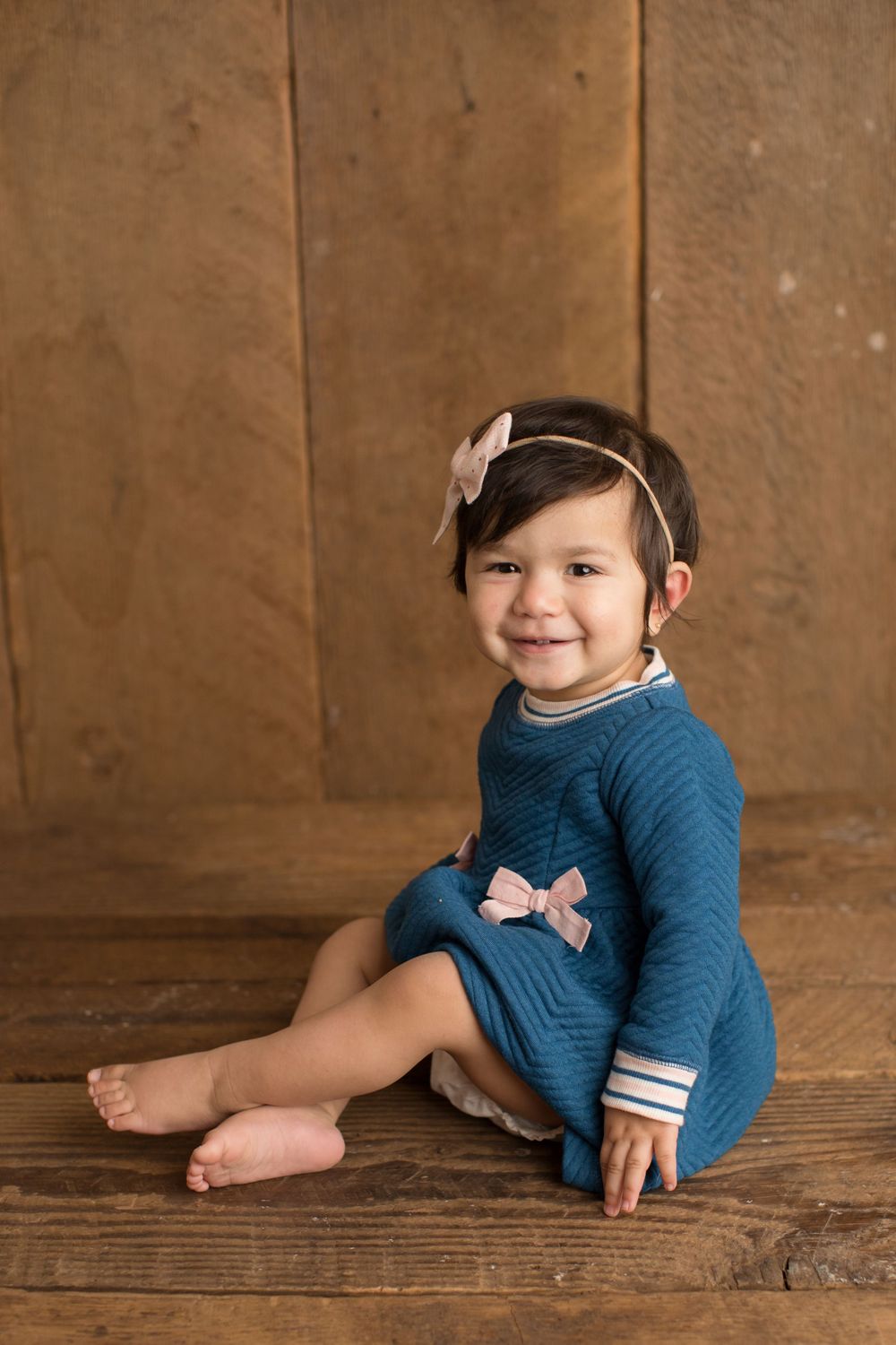 Child in blue dress sitting against wooden backdrop for portrait session.