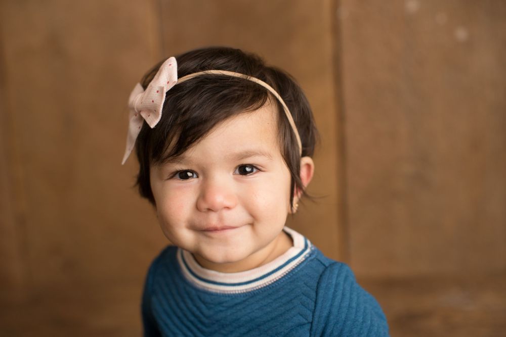 Small child wearing a blue sweater and bow headband smiling warmly against a brown wooden background.