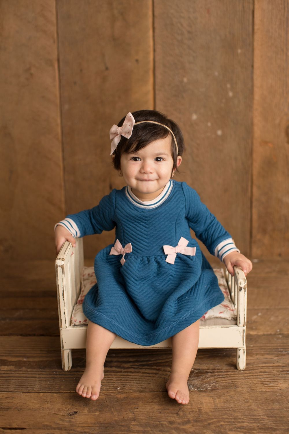 Child in blue dress with bow sits on vintage white bench against brown wooden wall.