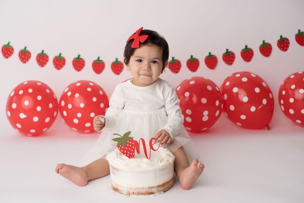 Baby in white dress with strawberry themed party decorations and cake.