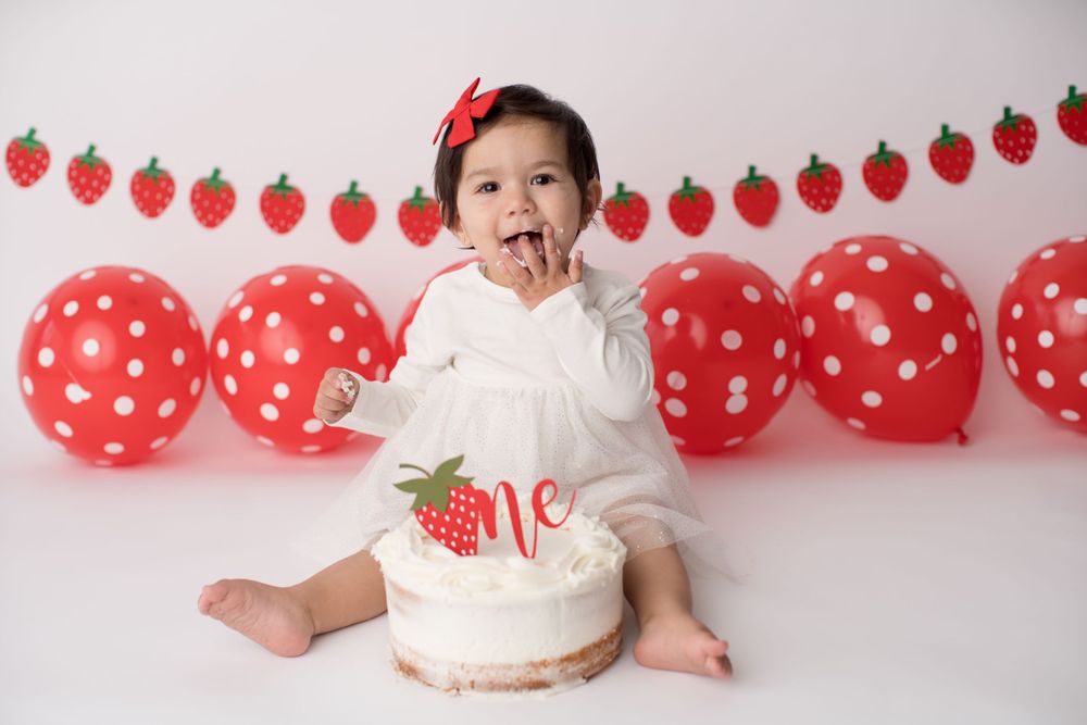 Baby celebrates with strawberry cake surrounded by red polka dot balloons and decorations.