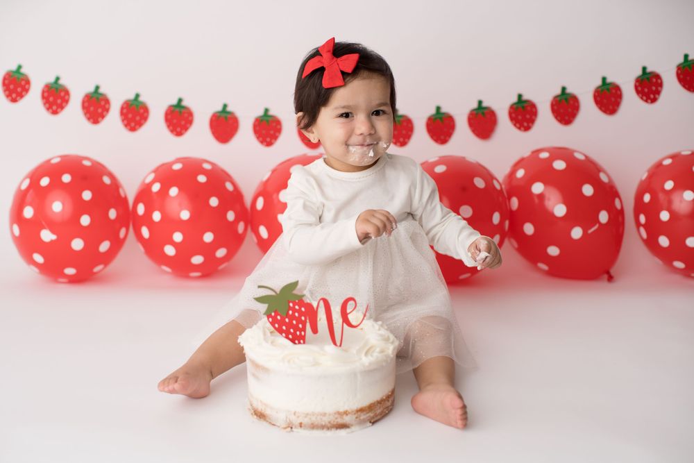 Baby in white dress enjoying strawberry themed cake with red balloon decorations.