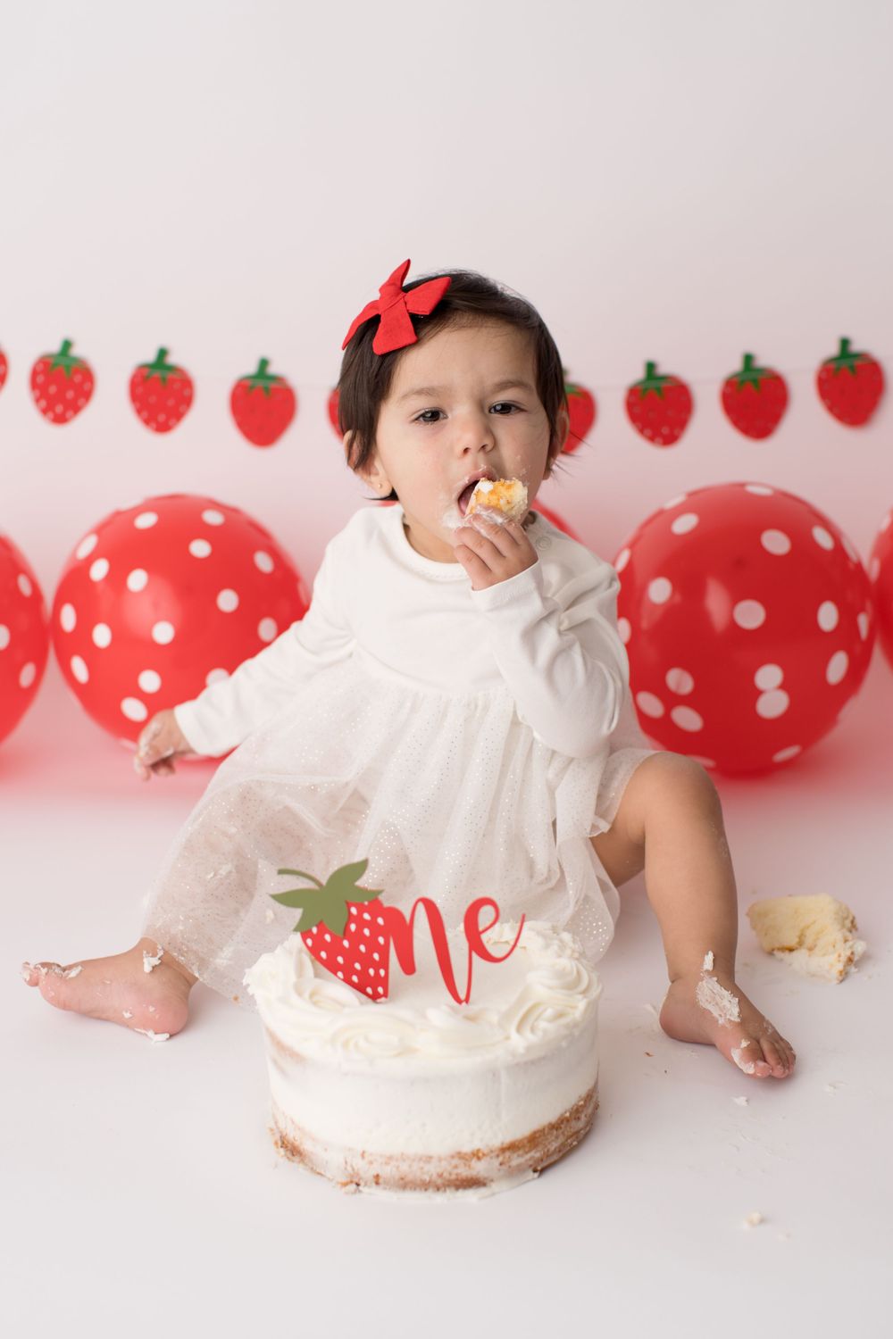 Baby eating cake at strawberry themed party with red balloon decorations.