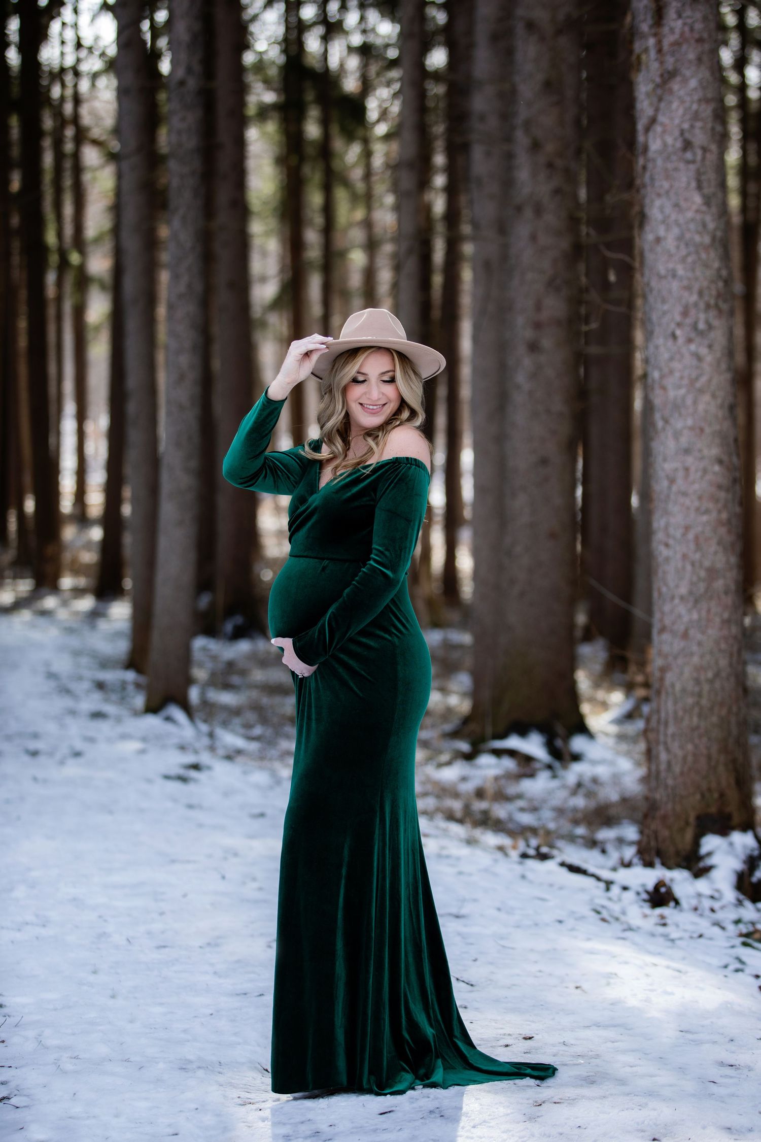 Maternity photo in an emerald velvet gown and hat amid snowy winter woods.