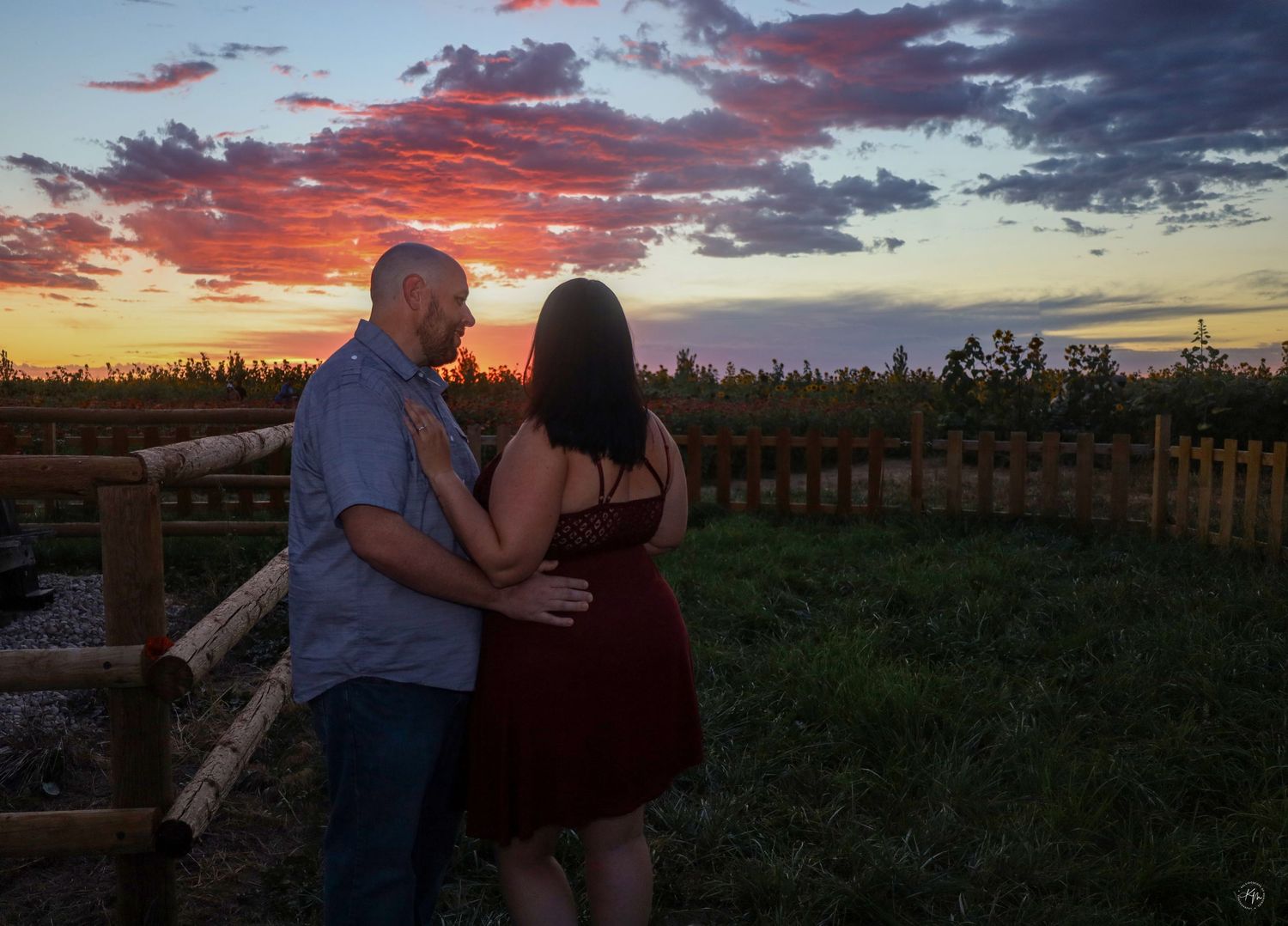 Romantic couple embracing while watching a dramatic sunset over a fenced rural landscape.