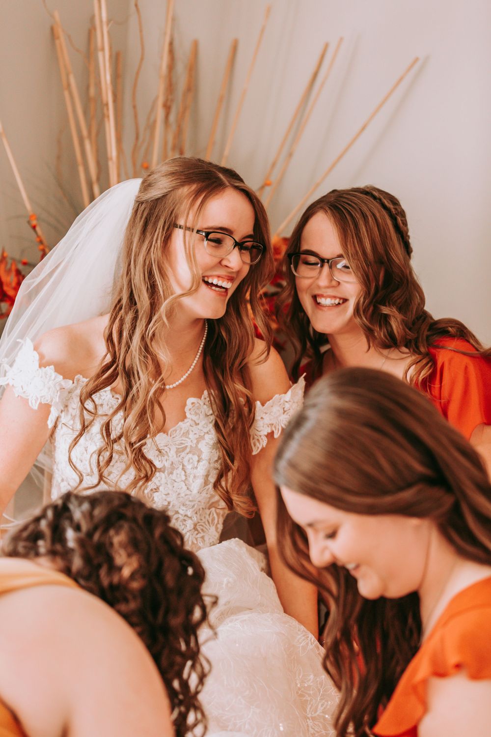 A bride in a lace dress and bridesmaids in orange share joyful laughter during a wedding celebration.