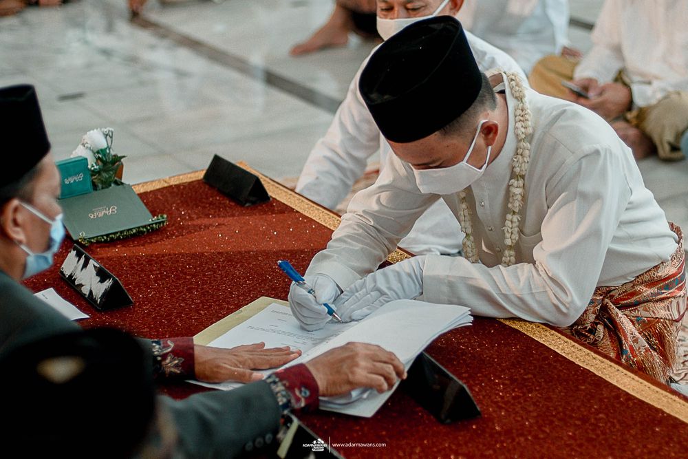 Wedding official in traditional dress signing ceremonial documents at red-covered table.