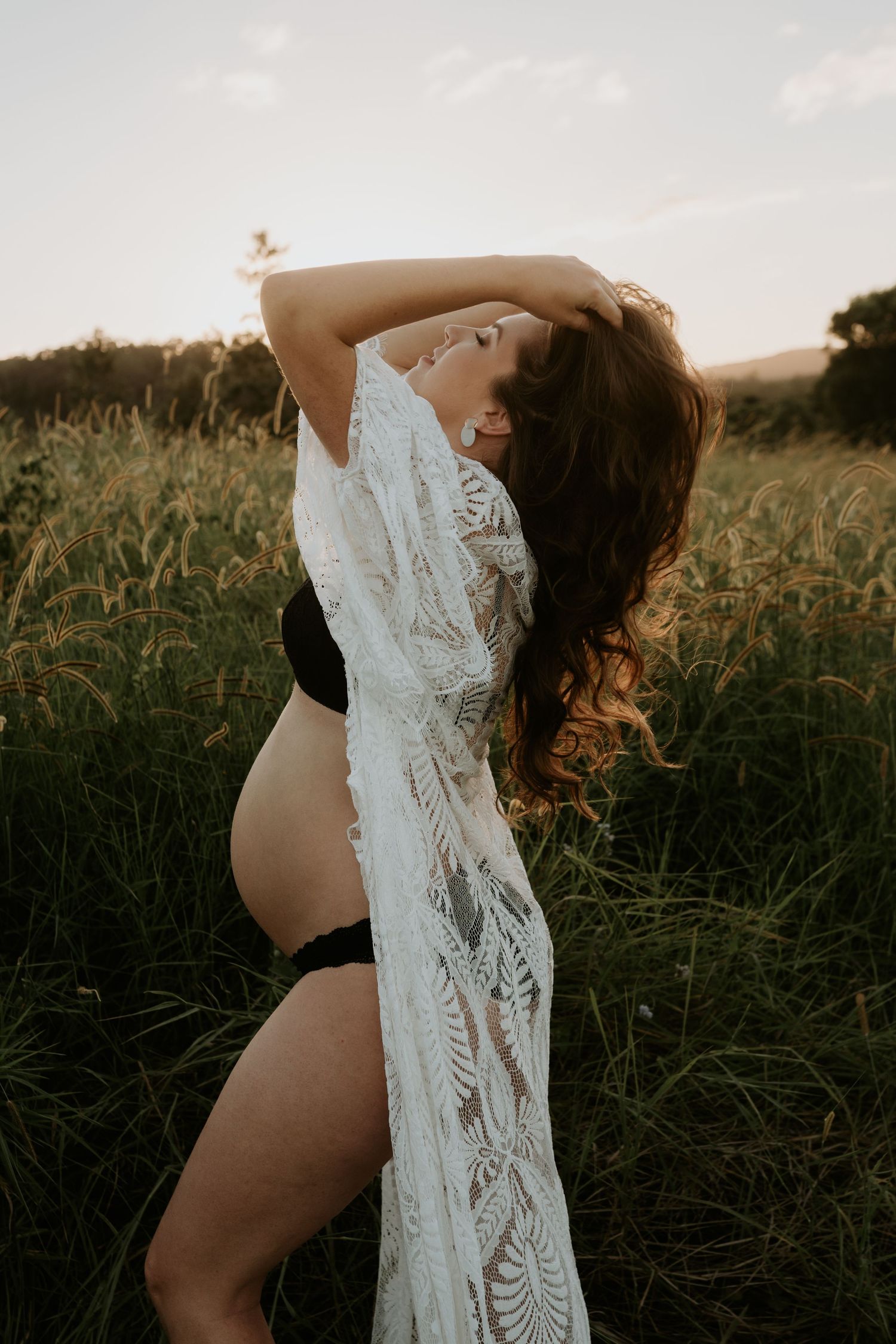 Artistic maternity silhouette of a woman in black lingerie holding white lace fabric at golden hour in a field.