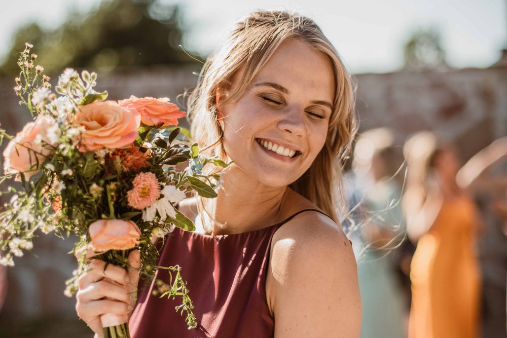 Person holding a vibrant bouquet of coral roses and wildflowers while smiling outdoors.