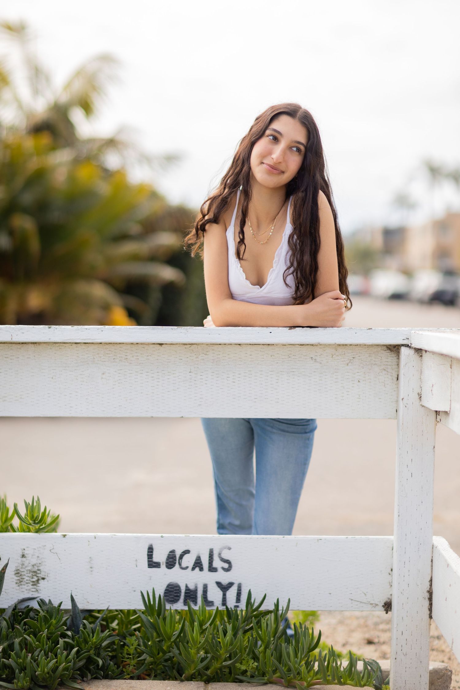 A high school senior in white top and jeans leaning on white fence with 'Locals Only' text painted on it at beach setting.