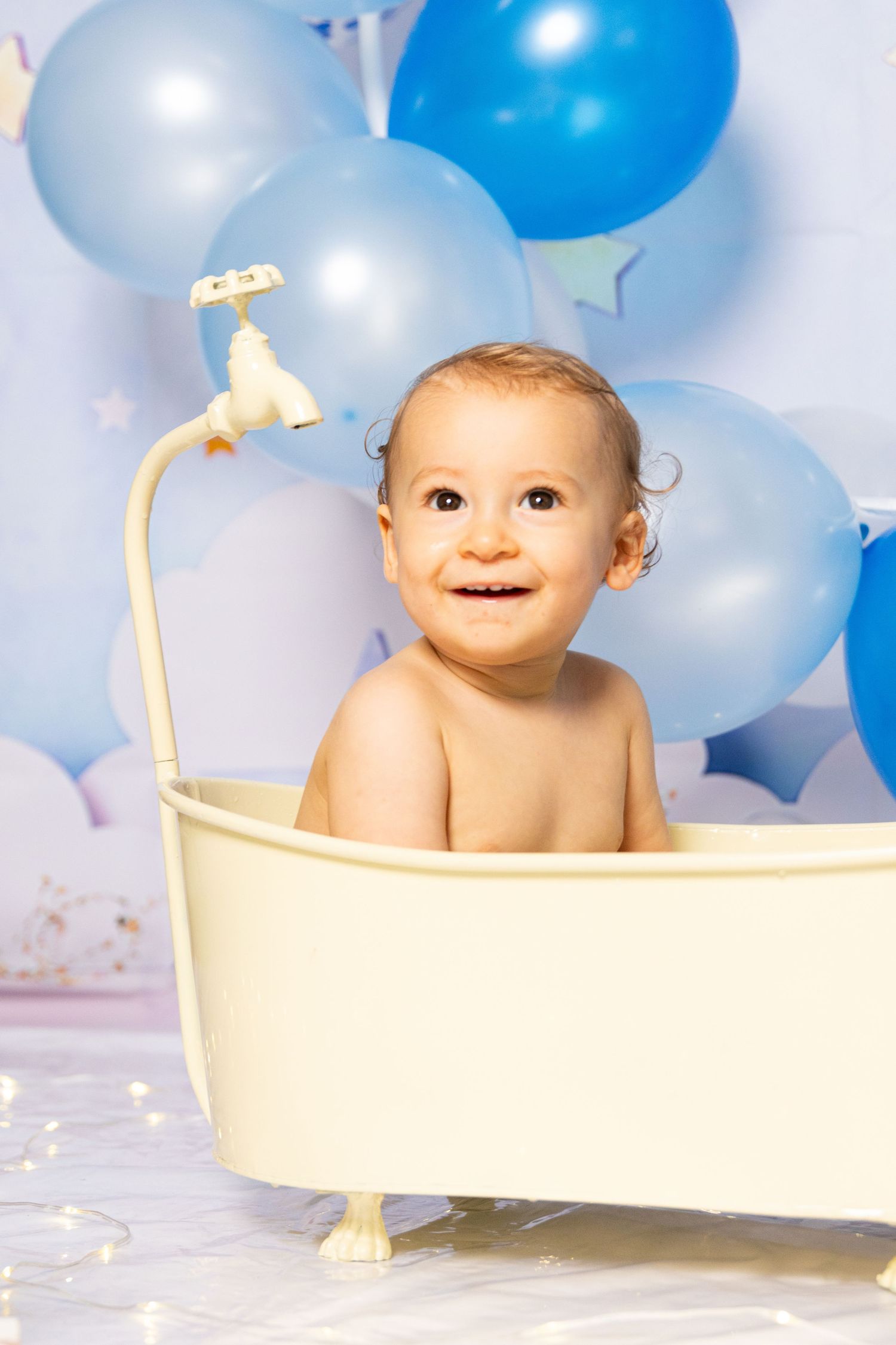 Baby sits in miniature bathtub prop with blue and white balloons in background.