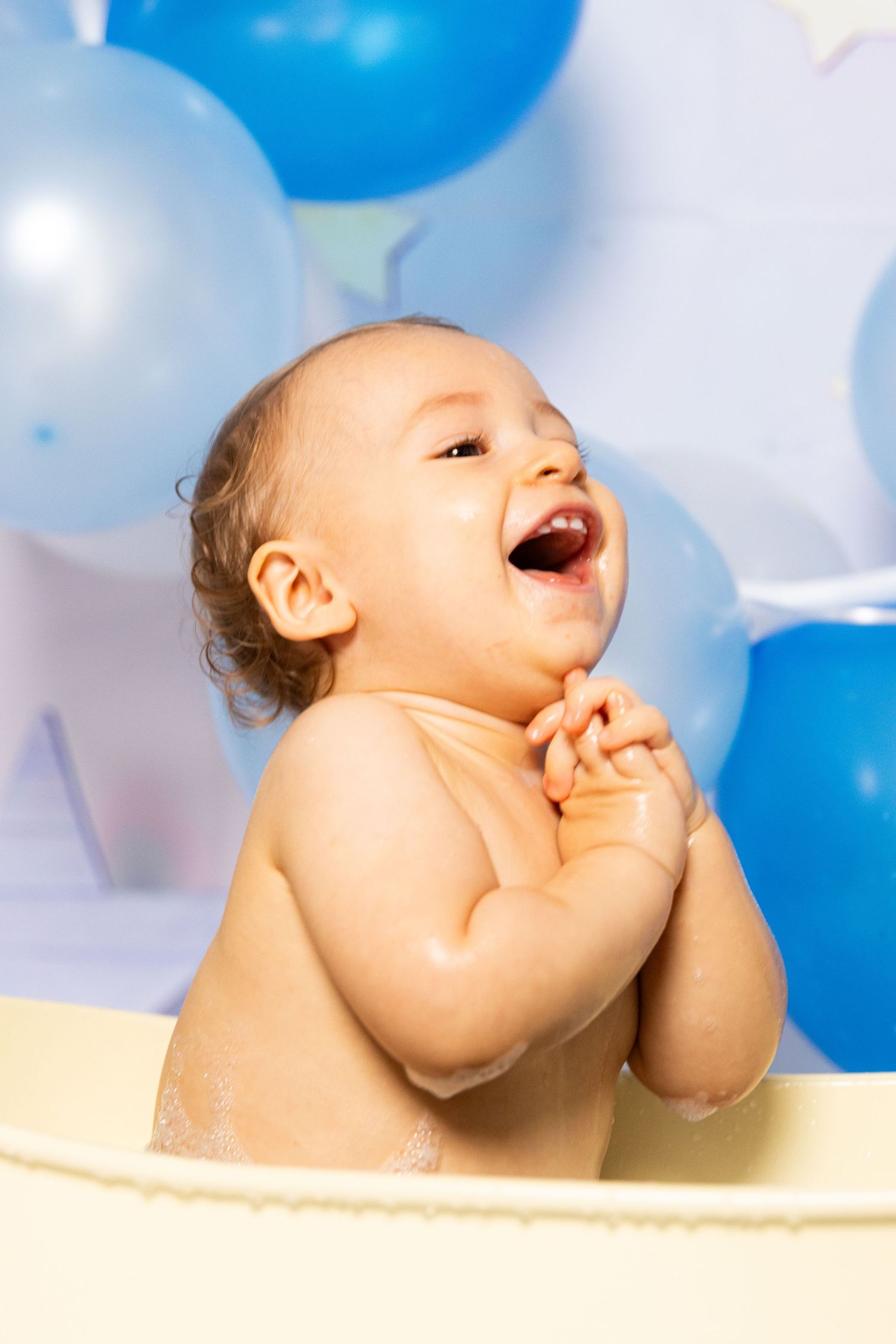 Laughing baby poses in cream-colored bath prop during photoshoot with blue balloons.