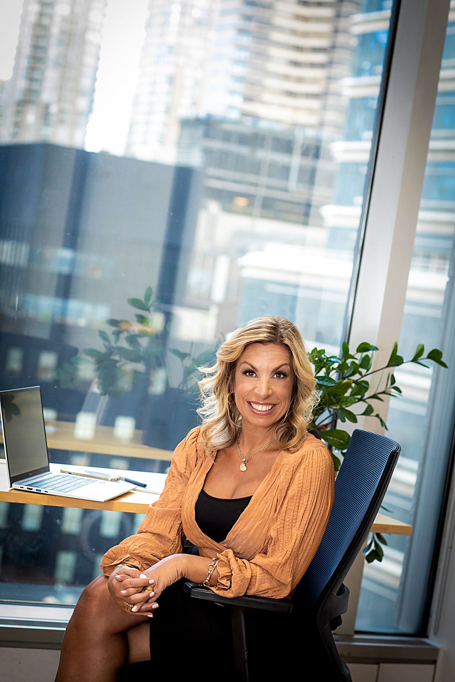 Professional seated in office chair wearing orange blouse against city skyline background.