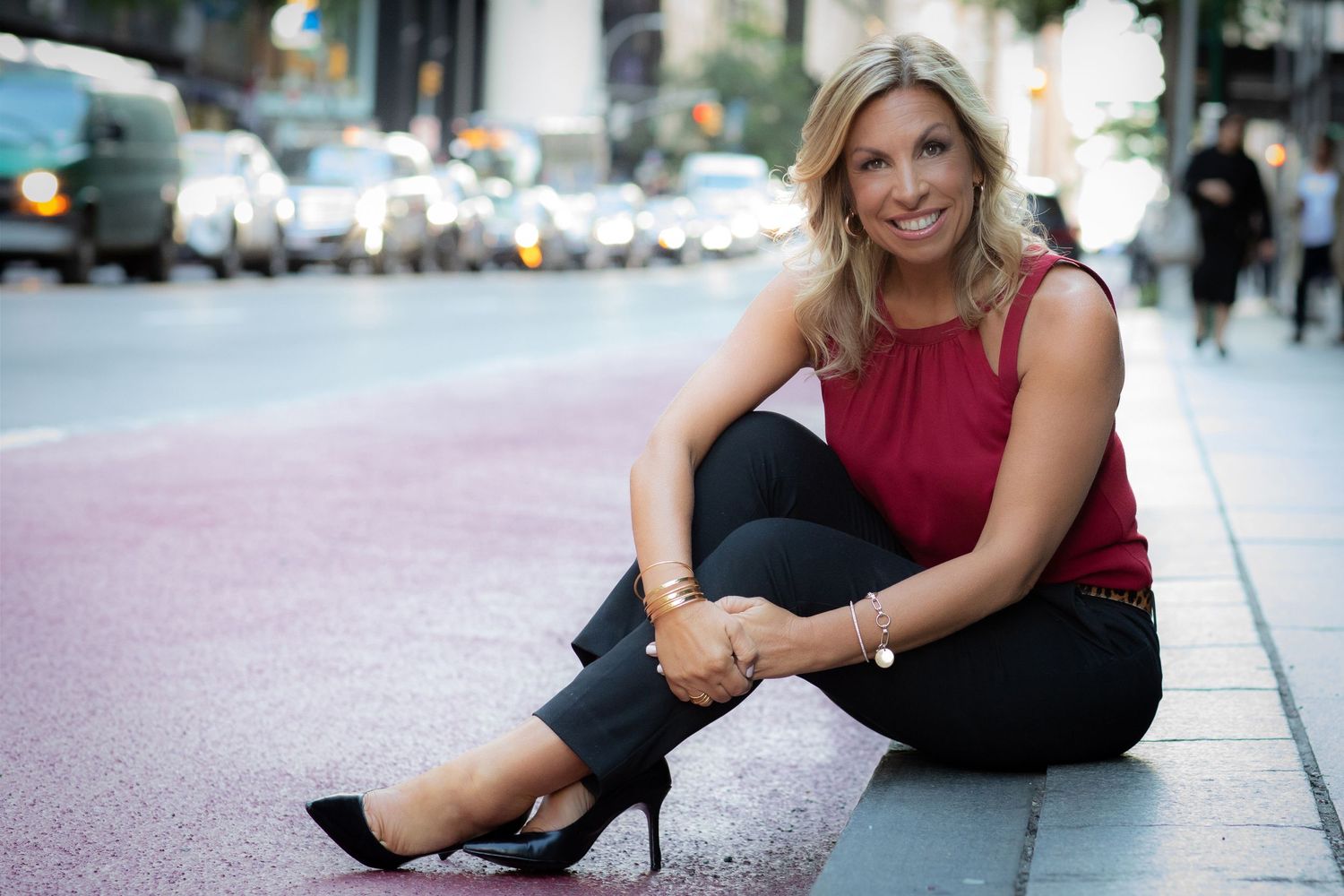 Casual outdoor portrait of person in red sleeveless top sitting on city sidewalk.