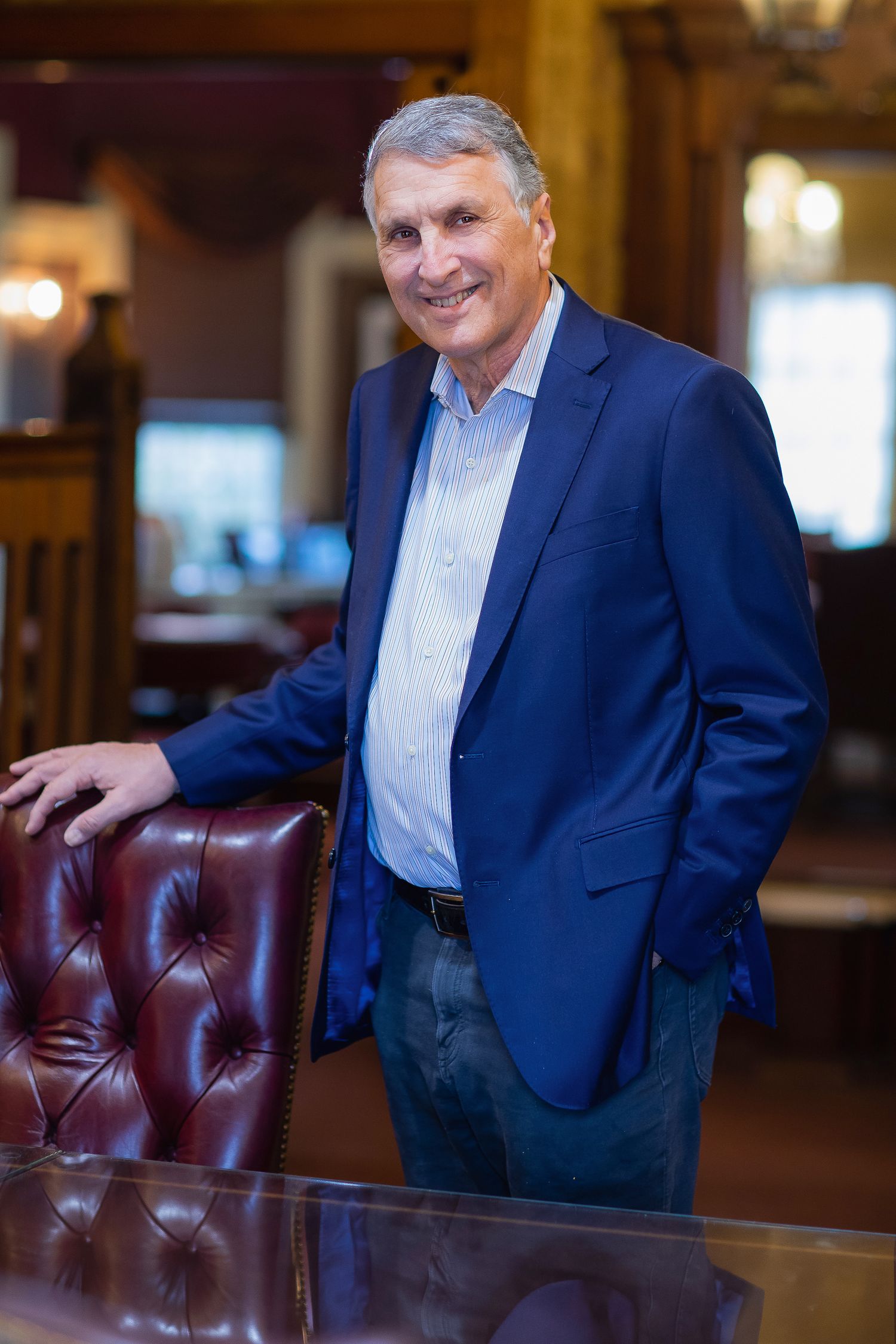 Executive in navy blazer standing next to leather chair in formal business setting.