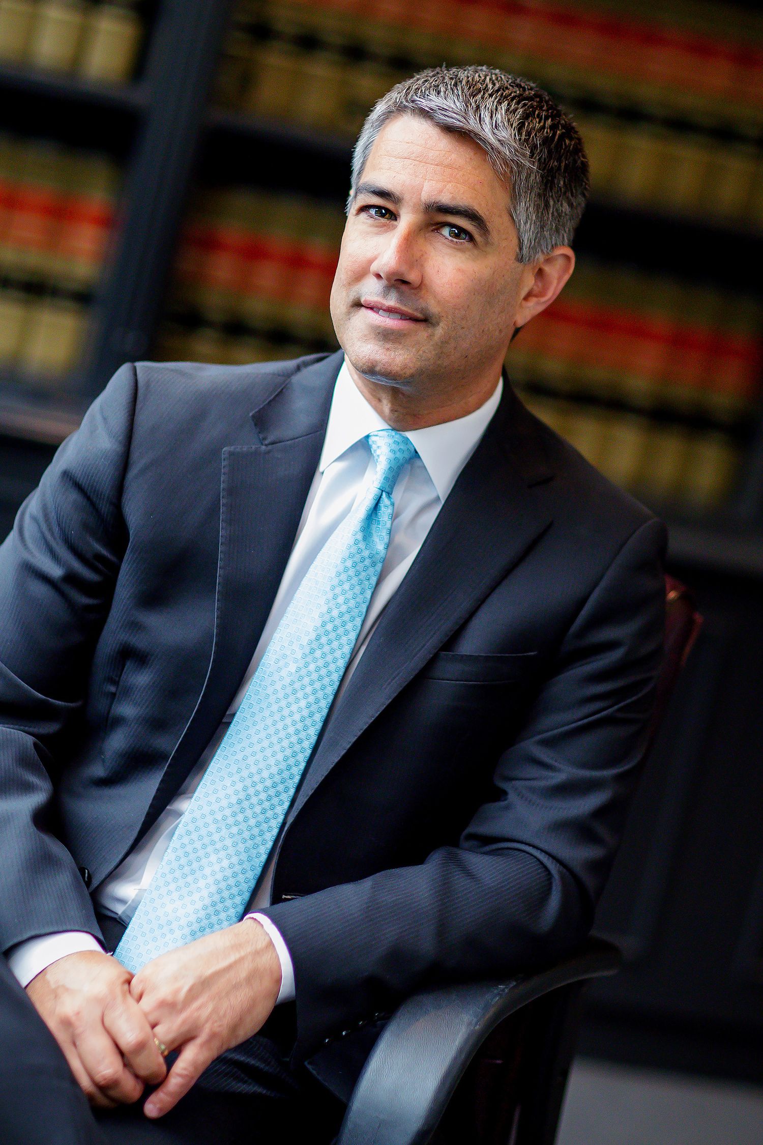 Professional headshot in navy suit and light blue tie against law library backdrop.