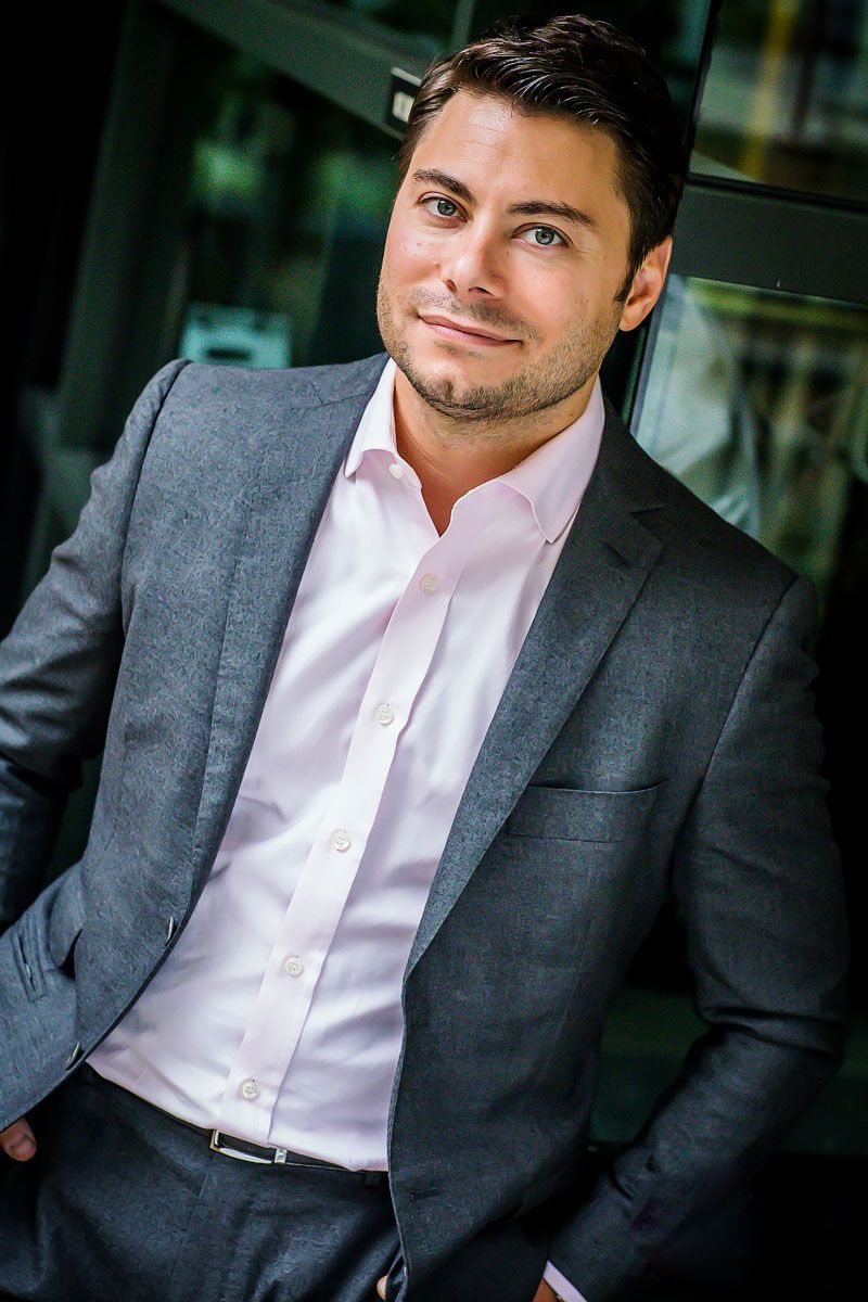 Professional portrait in grey suit and pink shirt against modern office background.