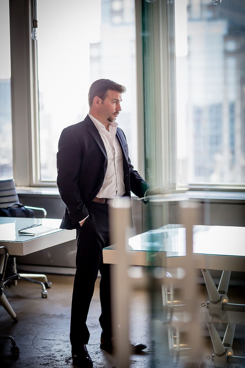 Professional in dark suit looking thoughtfully out office window in modern corporate setting.