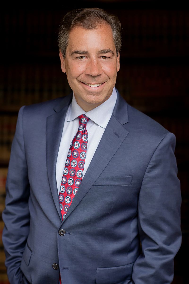 Professional headshot of person in blue suit and patterned tie against dark background.