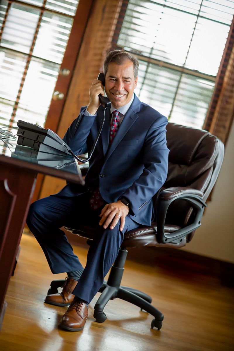 Business professional talking on phone while seated at desk in office with window blinds.