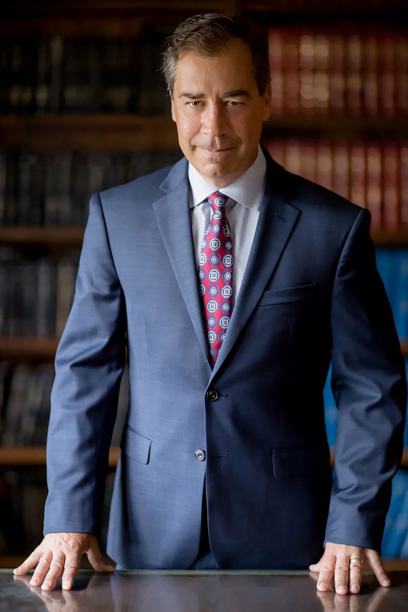 Professional standing at desk in blue suit with library bookshelf background.