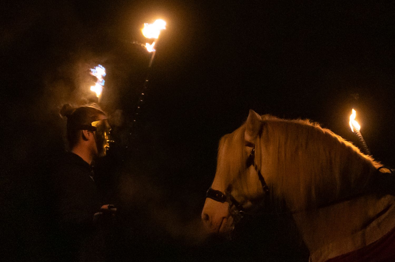 A masked performer holds flaming torches beside a white horse during a dramatic nighttime fire show.