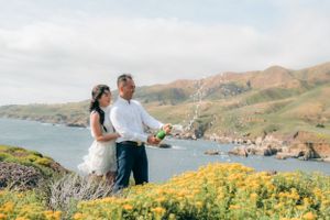 A couple stands on a coastal cliff overlooking the ocean with yellow wildflowers in the foreground.