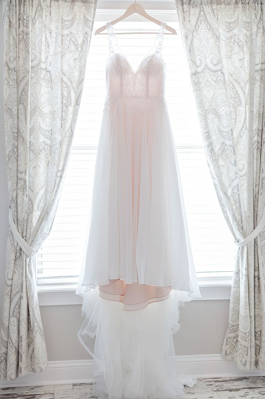 Wedding dresses hanging on hangers against lace curtains in a bright window.