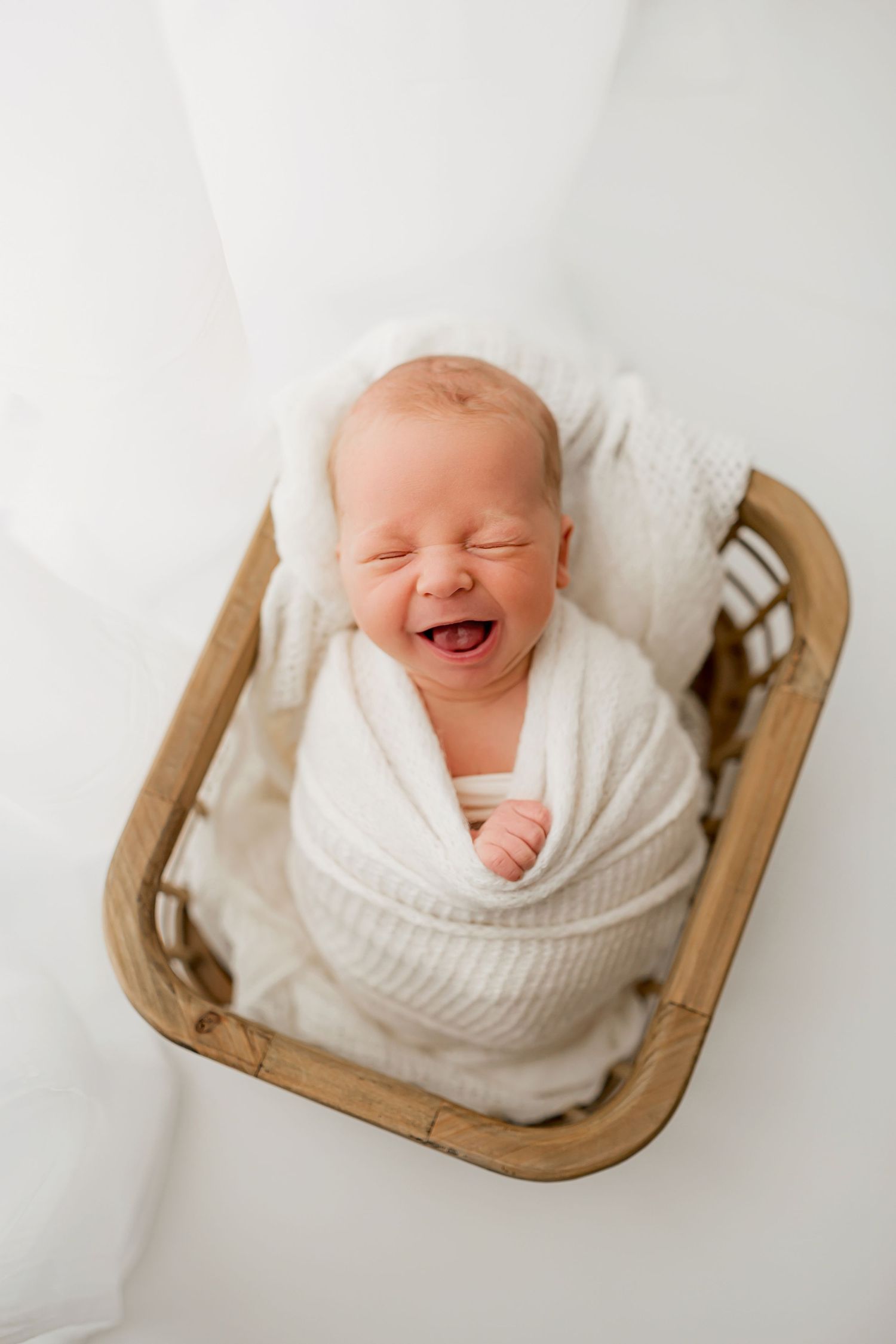 Baby boy laughing wrapped in white blanket in rattan basket in Flowertown Photography Studio in Summerville, SC