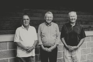 Three men stand together in casual attire against a brick wall in black and white.