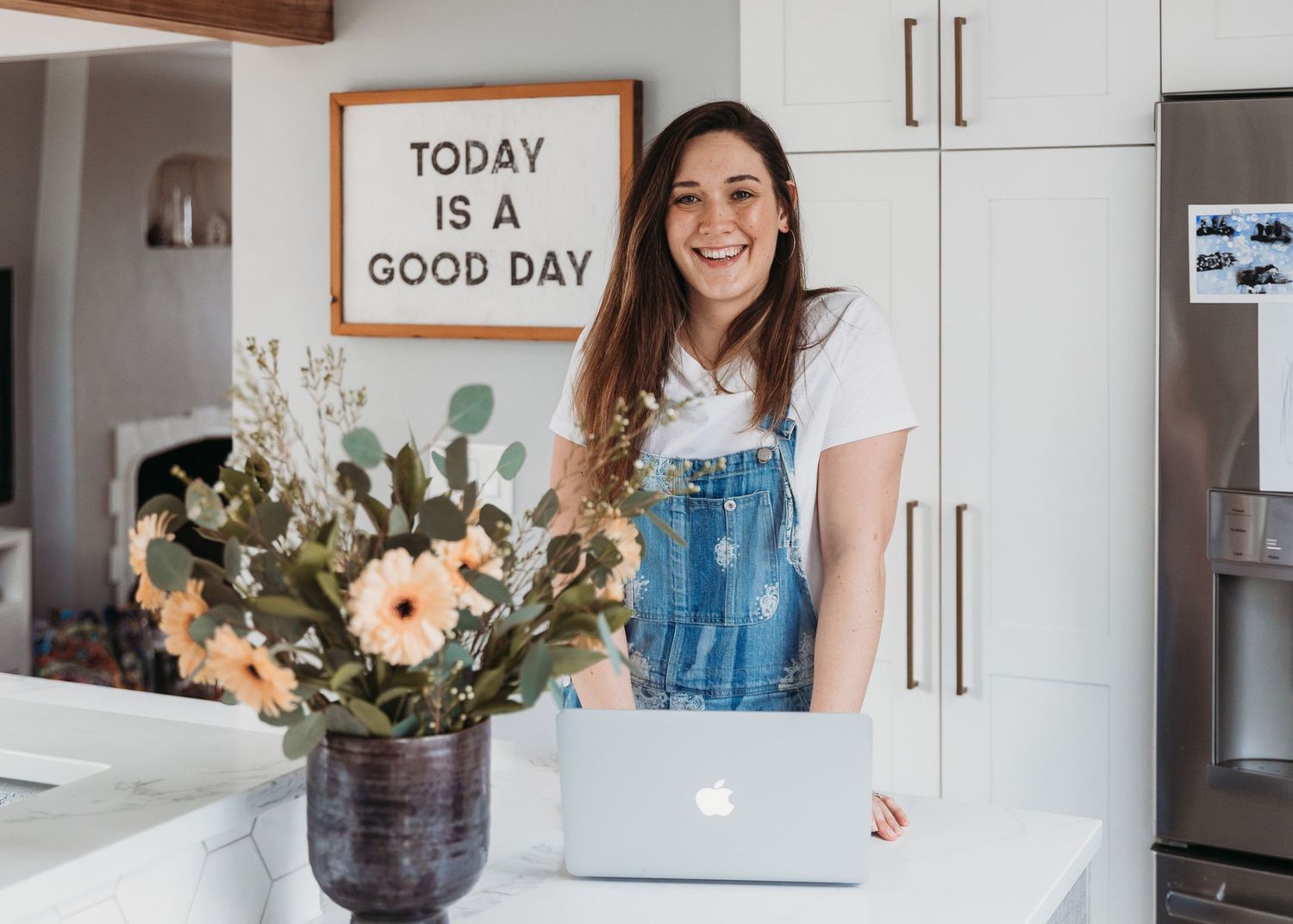 Woman in denim overalls working on laptop next to floral arrangement and motivational sign.