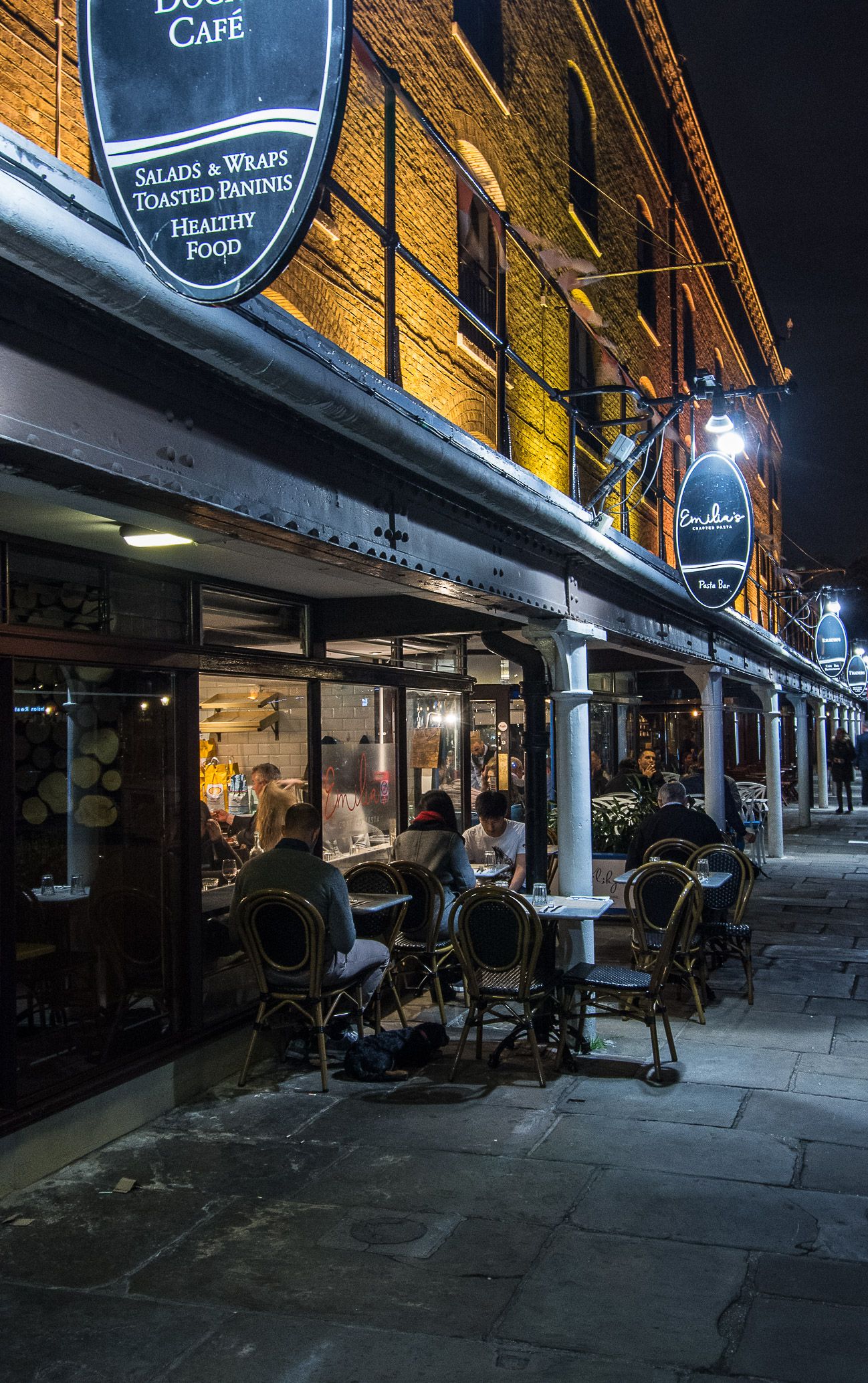 Cozy outdoor cafe seating area at night with illuminated storefronts and empty bistro chairs along a stone walkway.