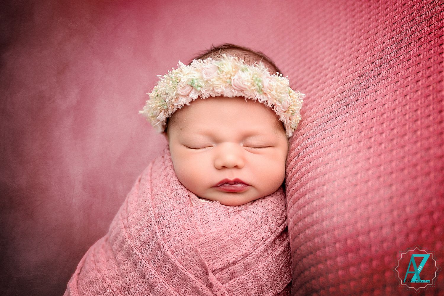 Baby wearing lace bonnet sleeps peacefully swaddled in pink wrap against rosy background.