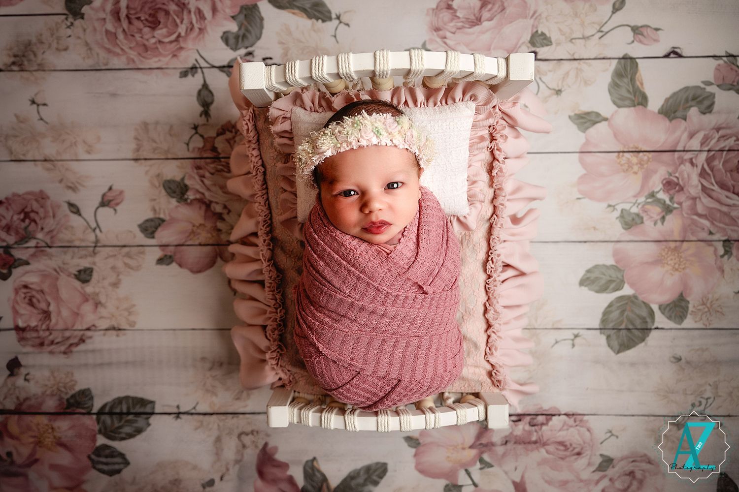 Newborn wrapped in pink swaddle rests on ruffled pillow against floral backdrop during professional photo session.