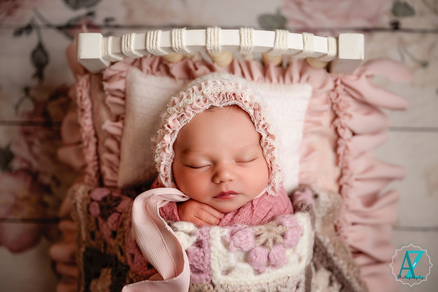 Sleeping newborn in vintage-style bonnet and pink wrap lies on decorative pillow with floral background.