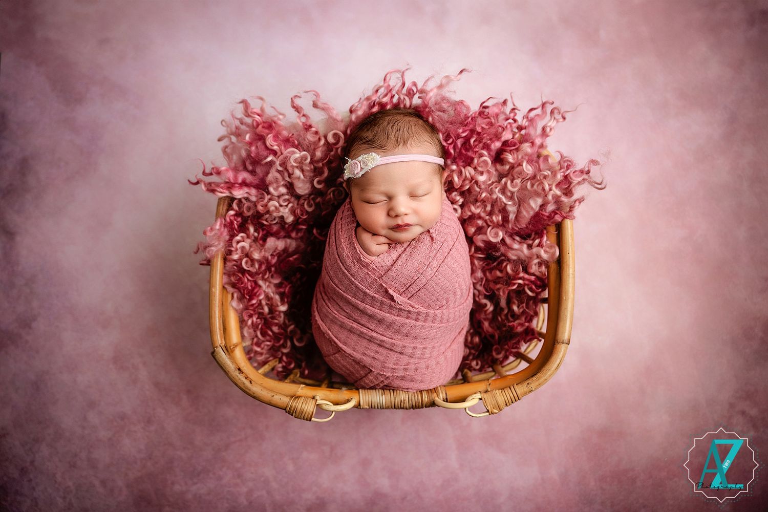 Sleeping baby wrapped in pink swaddle lies in vintage bassinet surrounded by pink floral decorations.