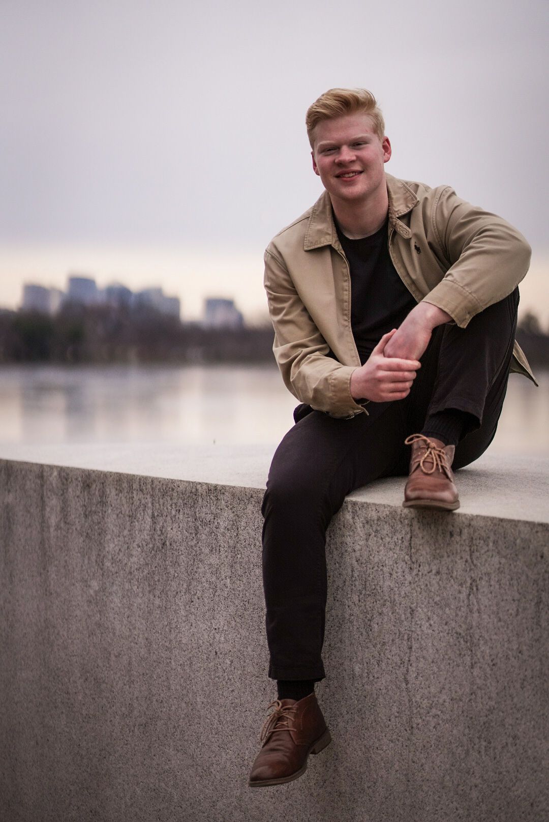 Person in tan jacket and dark pants sits casually on concrete wall with cityscape and water in background.