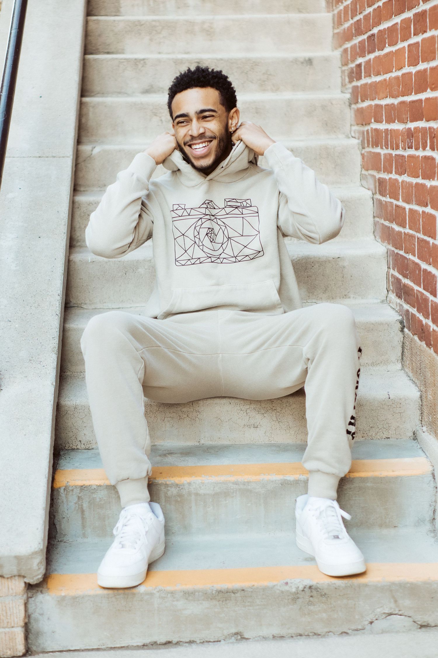 Person sitting on concrete steps wearing a casual beige hoodie with camera design and white sneakers.