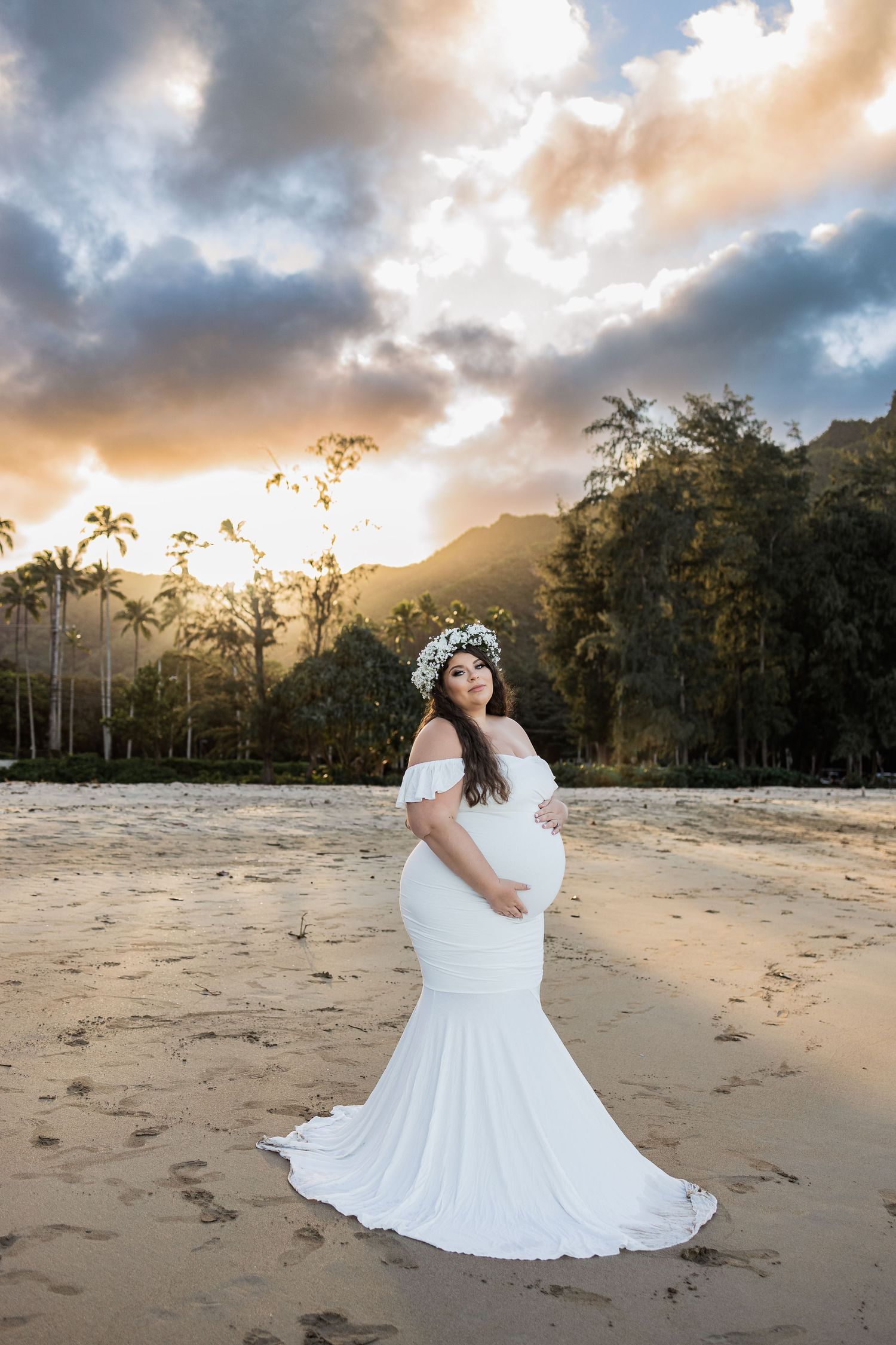 A maternity photo of a woman in a white dress on a Hawaiian beach during golden hour sunset.