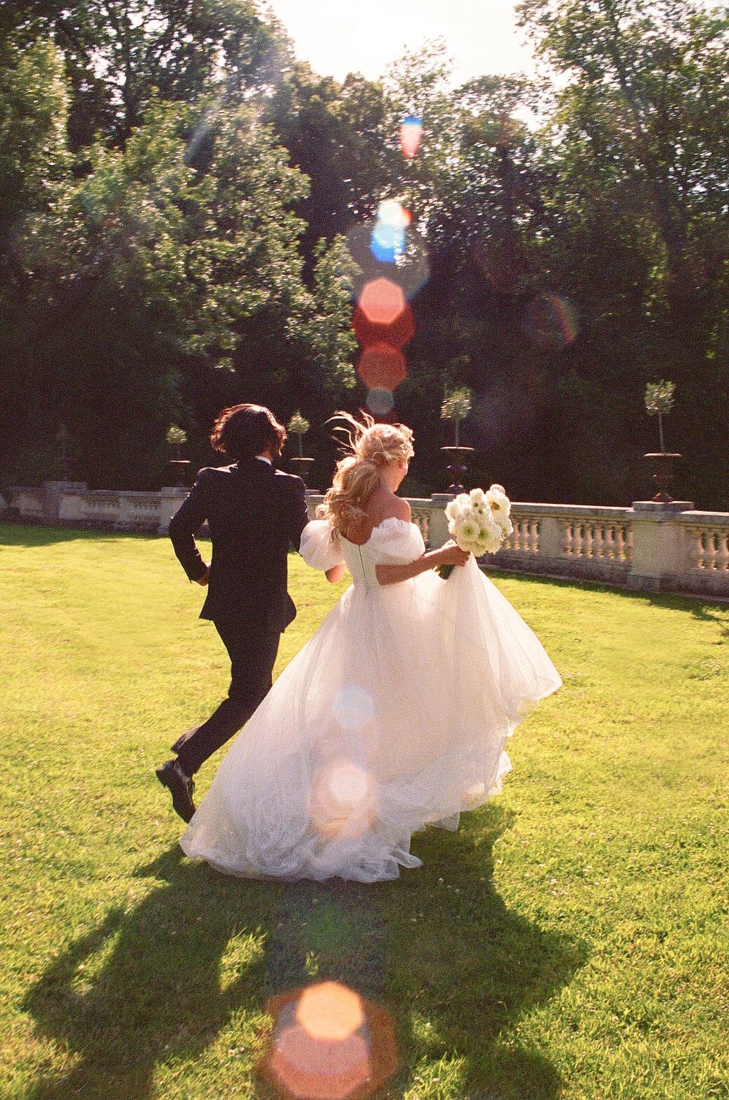 Newlyweds running joyfully across a sunlit garden lawn with lens flare effects.
