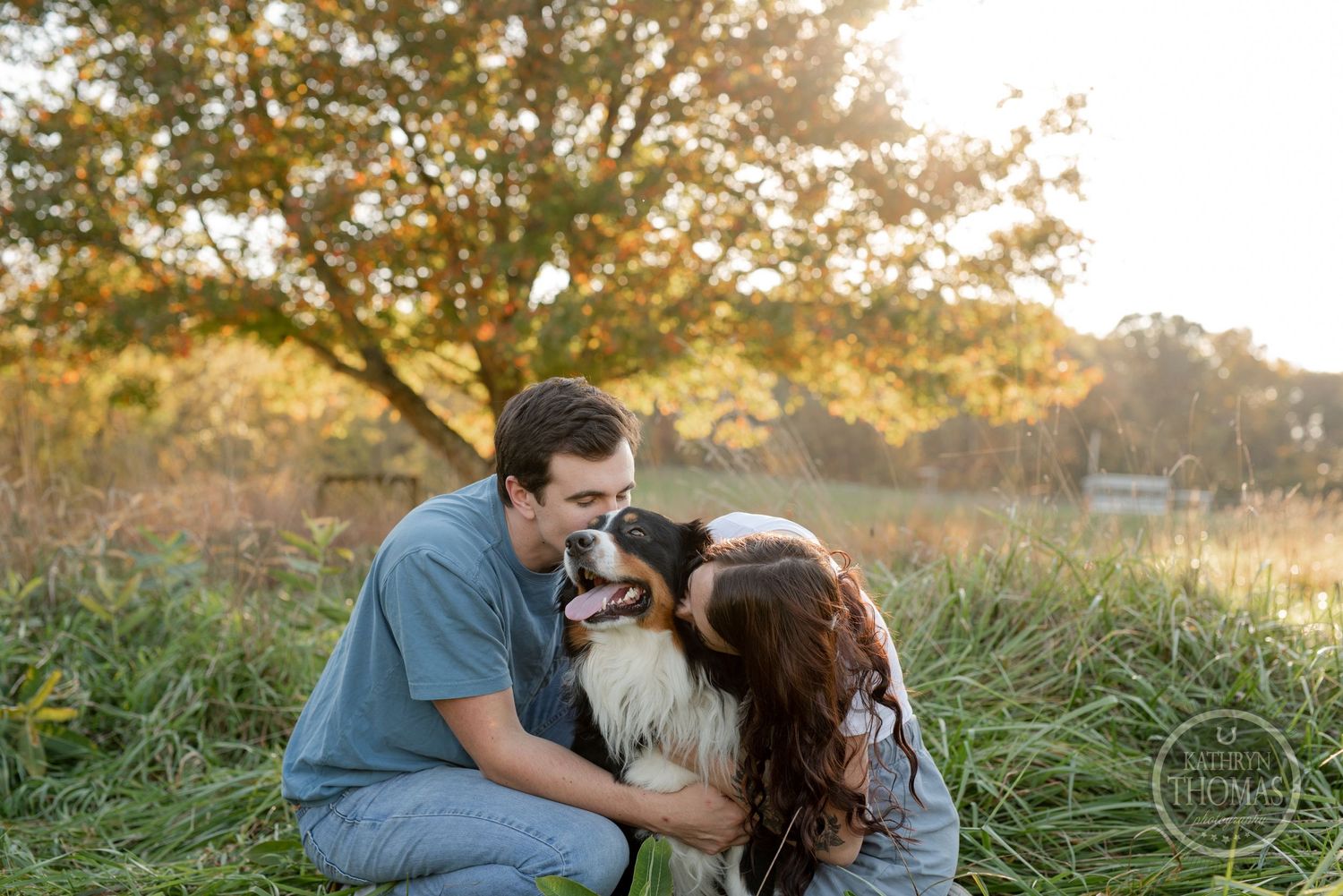 A couple pets their Border Collie while sitting in tall grass during sunset.