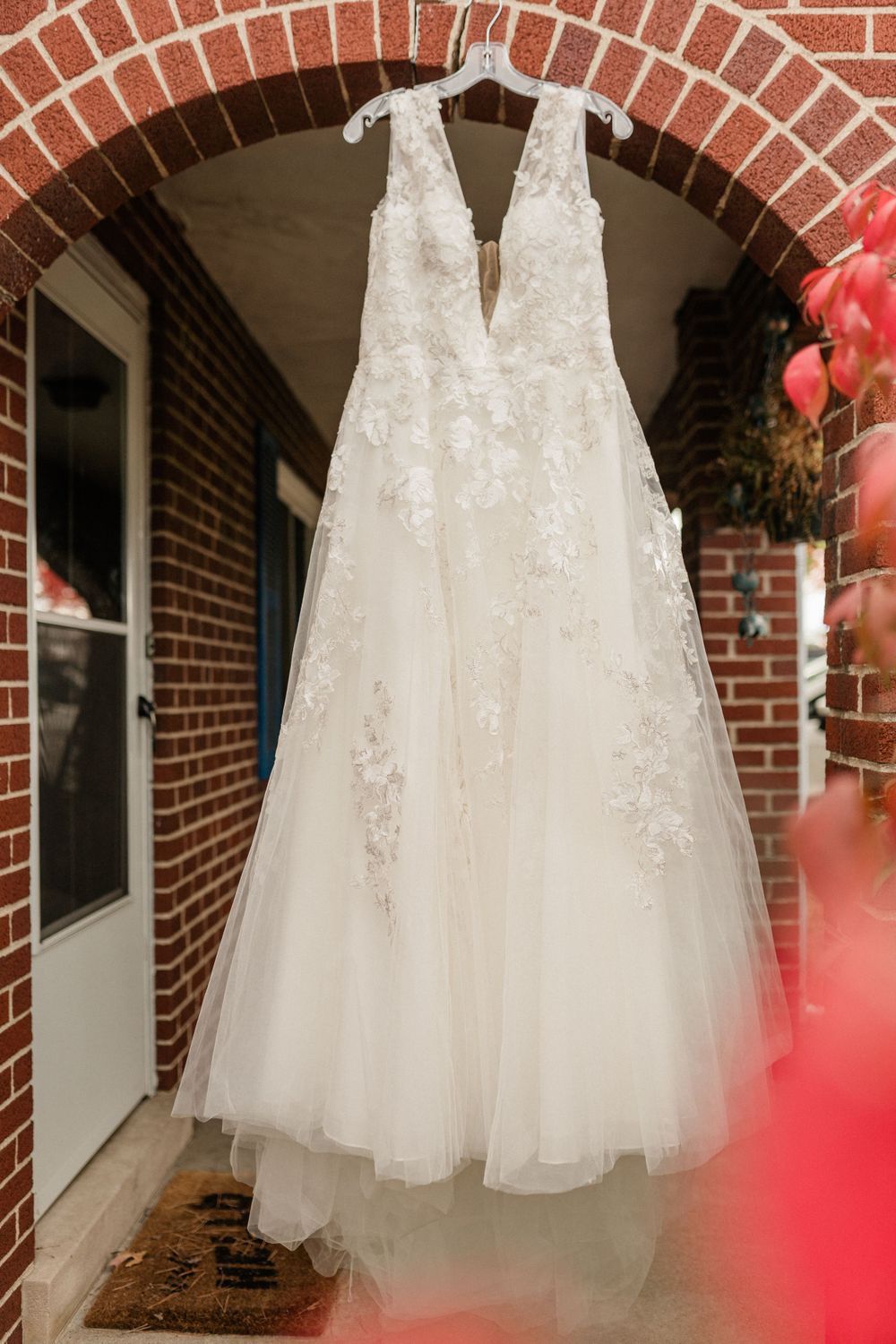 A white lace wedding gown with tulle skirt hangs in a brick archway.