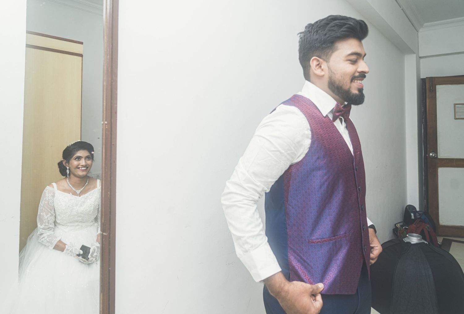 A groom in a purple vest and white shirt standing in a sunlit hallway.