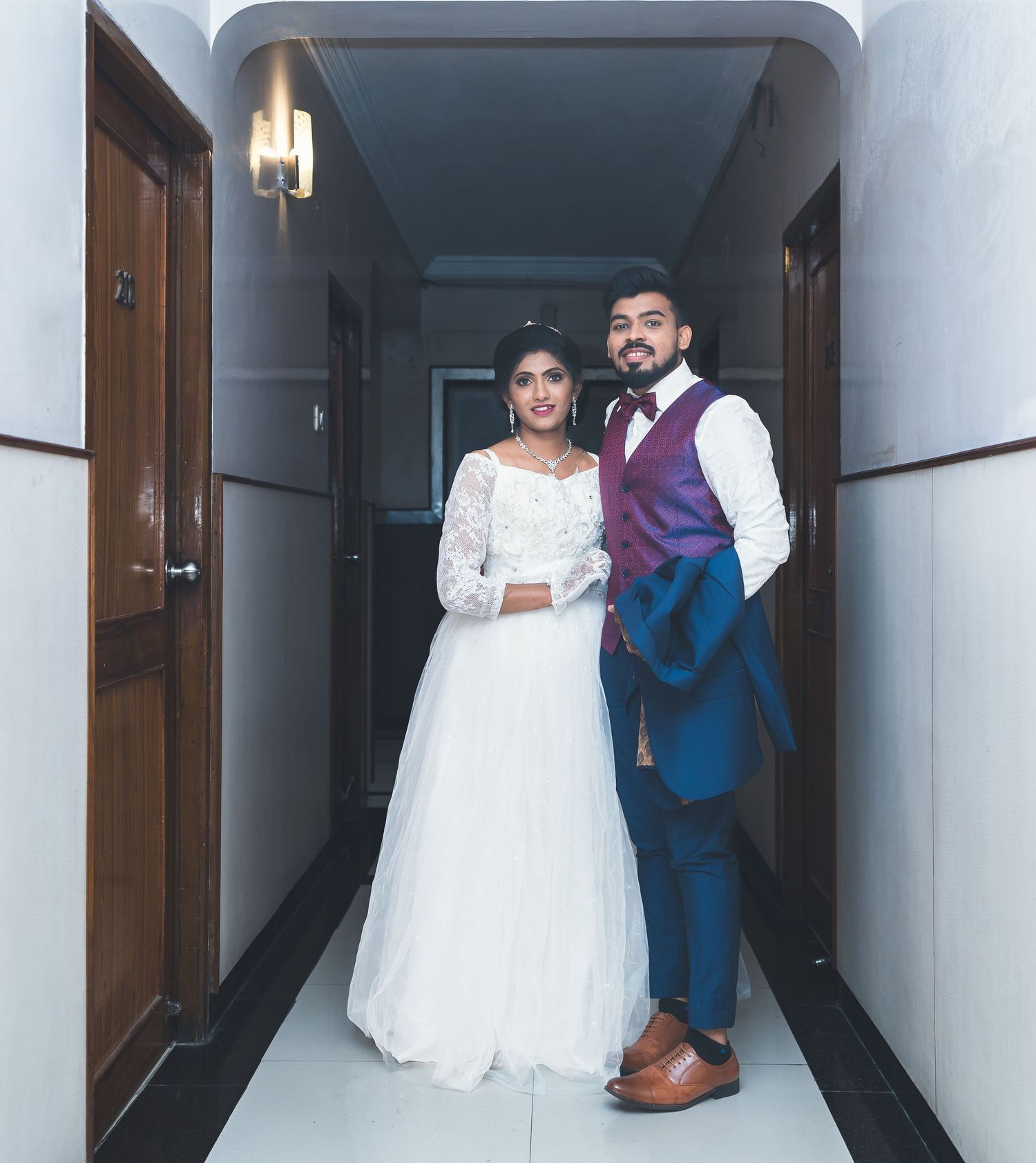 A newlywed couple poses in a hallway wearing elegant wedding attire.