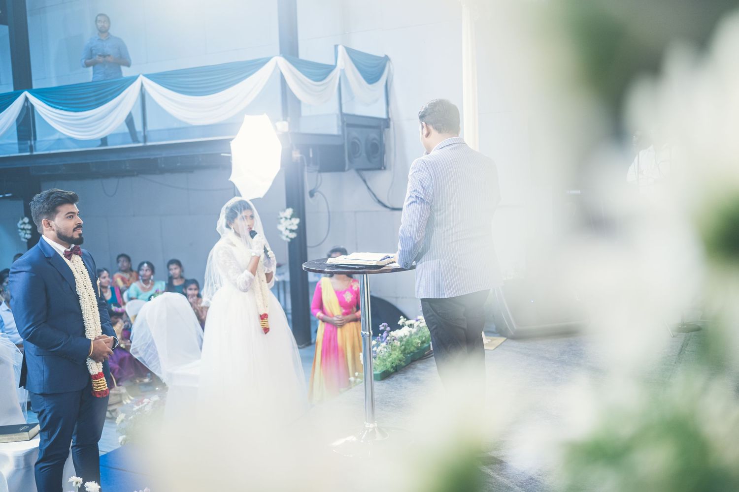 Wedding ceremony taking place under blue and white draped decor with guests seated in rows.