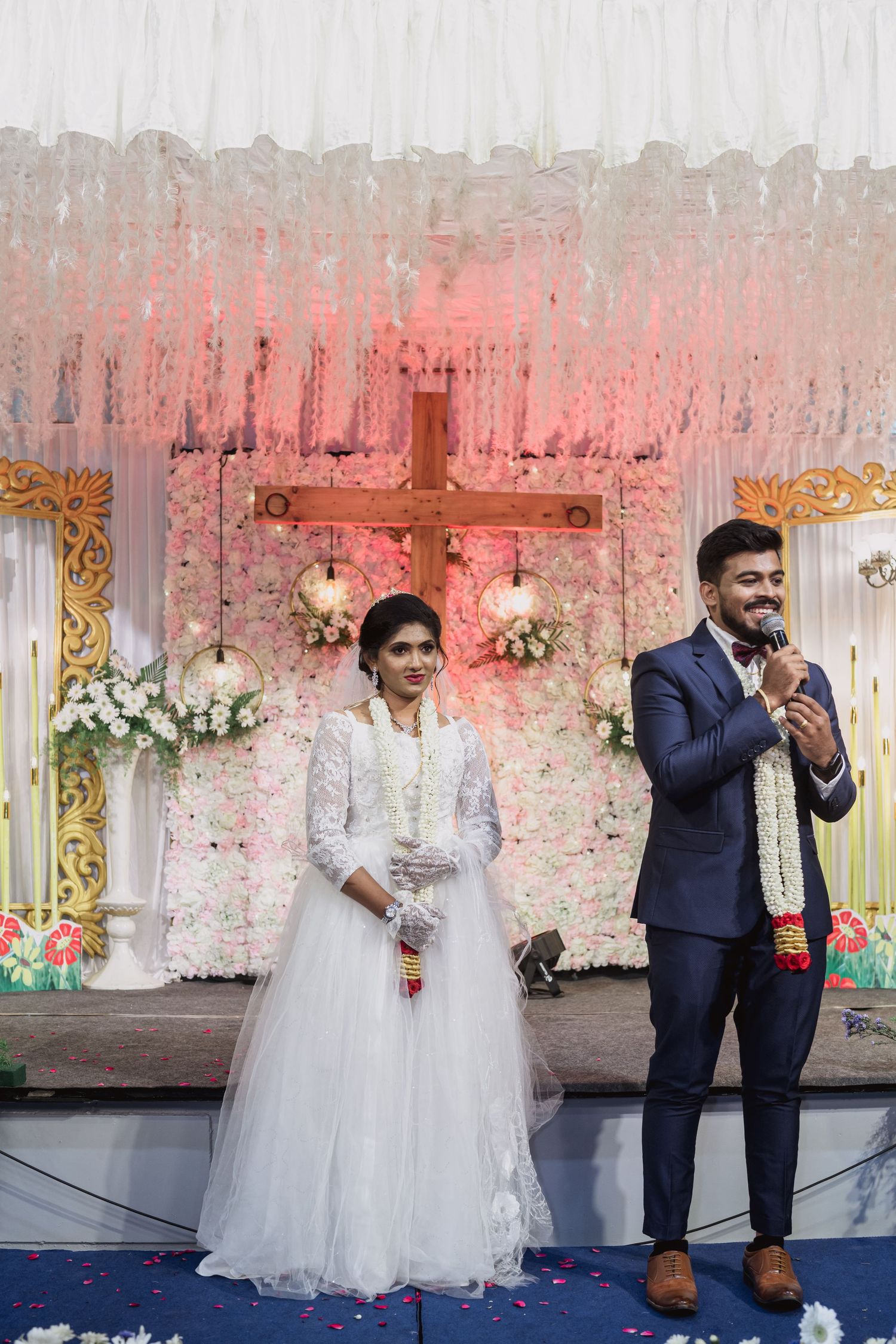 Wedding couple stands on stage with illuminated cross and floral backdrop during ceremony.
