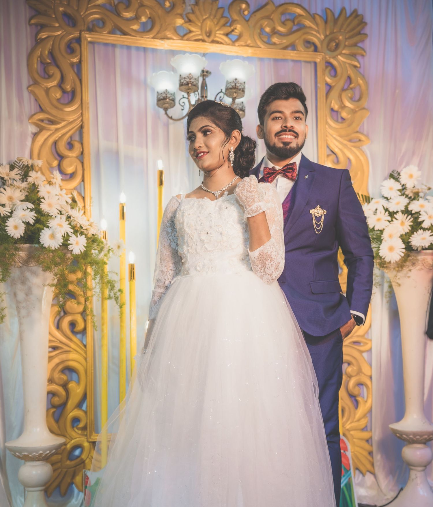 A couple poses in elegant wedding attire against an ornate gold frame and floral decorations.