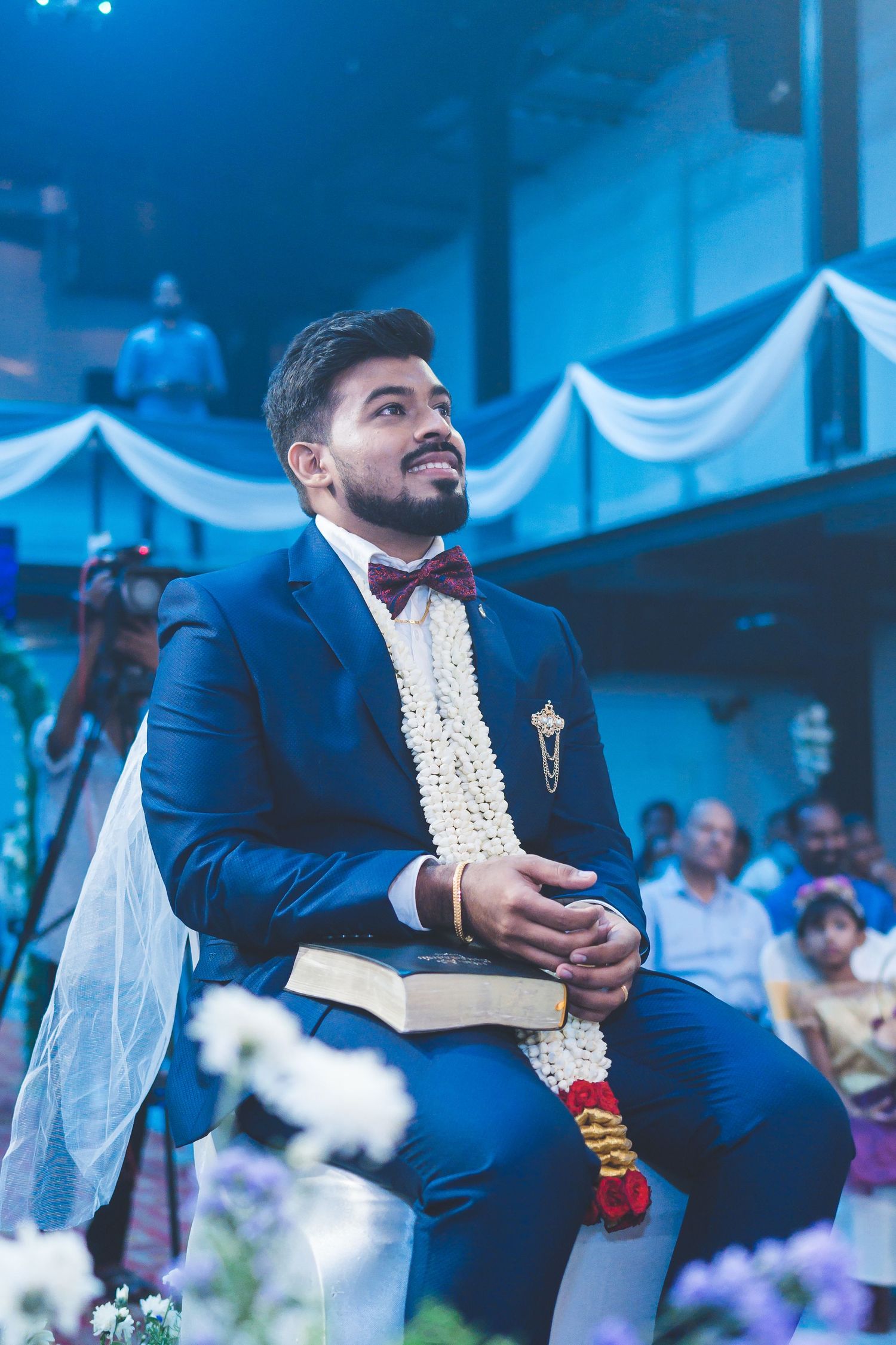 Groom wearing navy blue suit and bow tie with traditional white garland during ceremony.