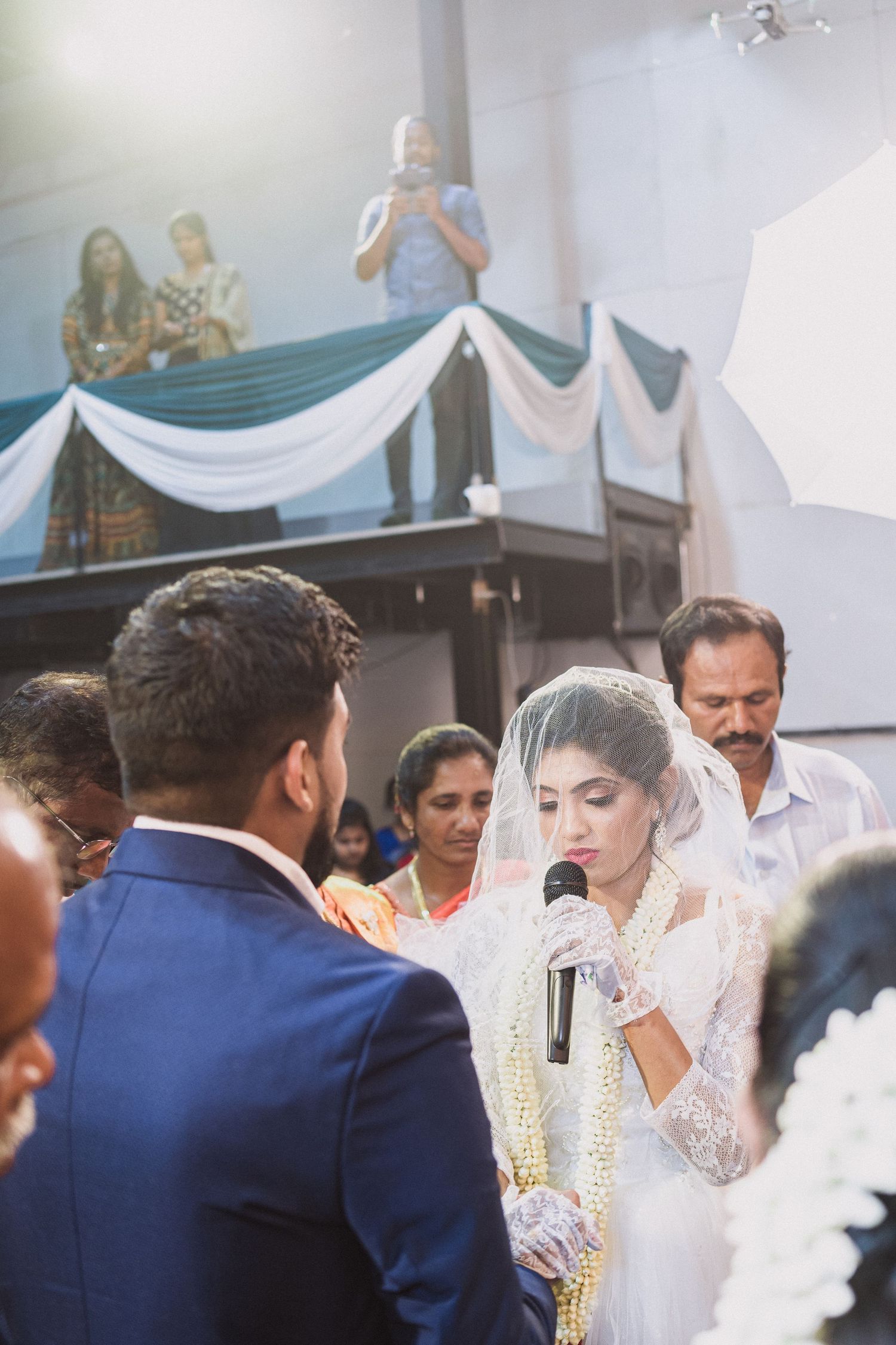 Indoor wedding ceremony with white draped decorations and guests watching from elevated seating.