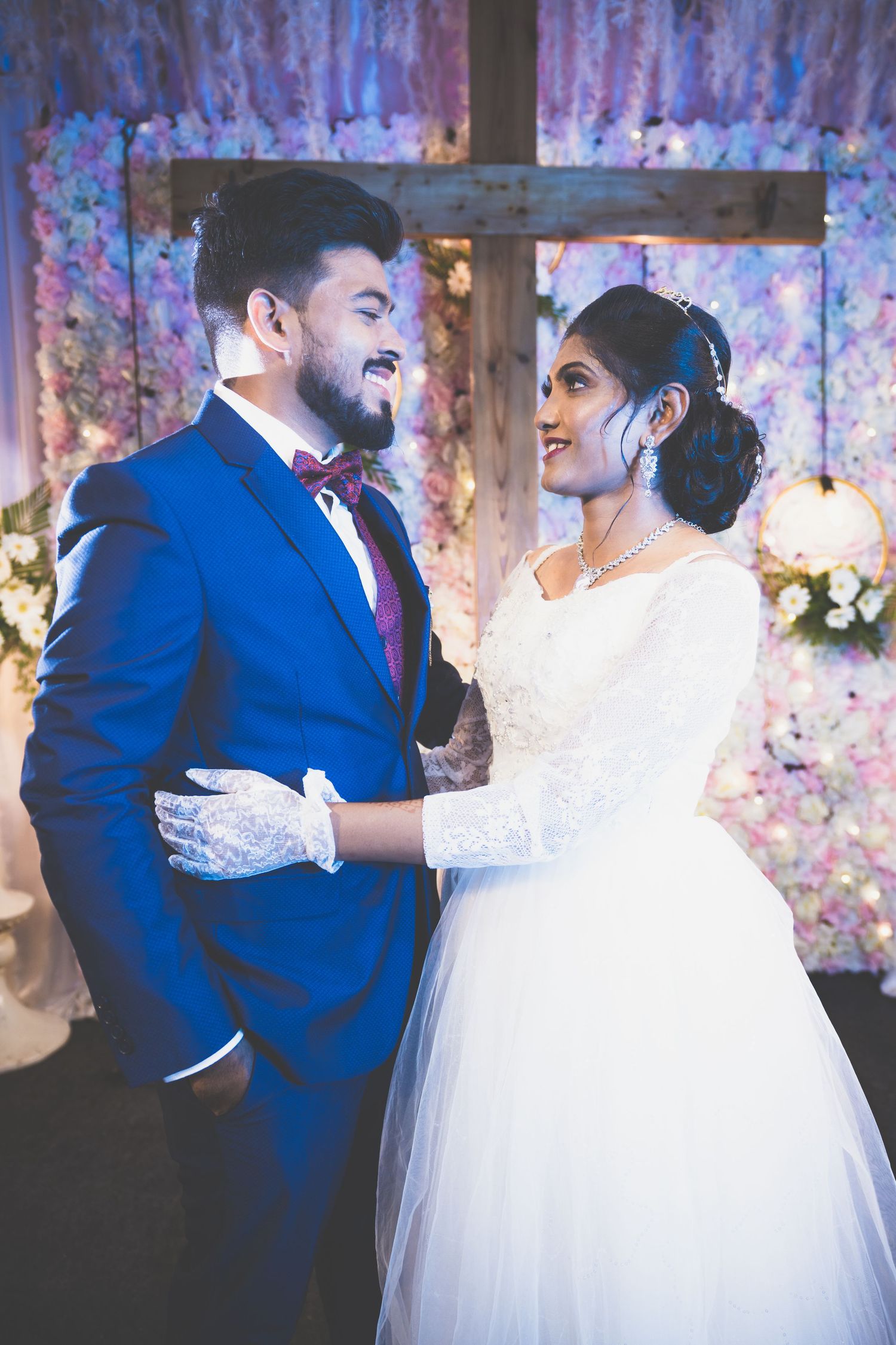A newlywed couple shares a joyful moment in front of a floral backdrop with a wooden cross.